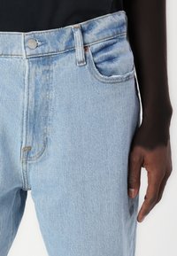 Los jeans de mezclilla azul claro presentan un diseño estándar de cinco bolsillos, cierre de cremallera y herrajes plateados, con un ajuste relajado y una textura sutil.