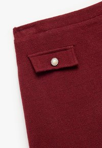 Tejido texturizado burdeos con un bolsillo de solapa que presenta un botón adornado con perla y detalles en plata. La vista en primer plano resalta el detalle del material.