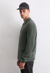 Groene geribbelde cardigan met een knoopsluiting aan de voorkant, lange mouwen en een platte kraag. Draagt over een zwart shirt en gecombineerd met donker gemusterde broek.