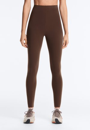 Femeie purtând colanți maro cu talie înaltă și pantofi sport albi cu șireturi gri, stând în picioare pe un fundal simplu, deschis la culoare.