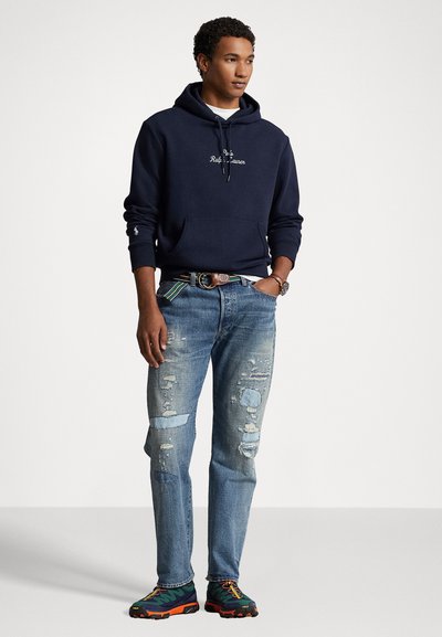 Jūrinio stiliaus sweatshirtas su priekinėmis kišenėmis, baltu logotipu, dėvėti šviesiai mėlyni džinsai ir daugiaspalviai sportiniai batai su tekstūruota paviršiumi ir ryškiais padais.