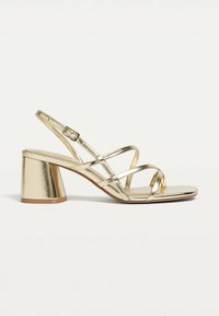 Gold metallic block heel sandal with thin crisscross straps and ankle buckle, displayed on a white background.