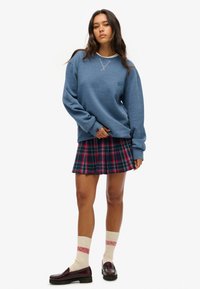 Sudadera azul con cuello redondo, falda de cuadros en colores oscuros, calcetines beige con rayas rojas y mocasines negros brillantes.