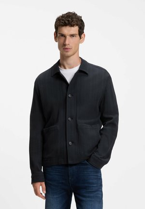Mann trägt dunkel geknöpfte Jacke mit Fronttaschen über weißem Hemd und blauen Jeans, steht mit einer Hand in der Tasche vor weißem Hintergrund.