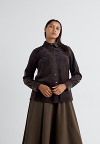 Donkerbruine corduroy blouse met een kraag, knoopsluiting aan de voorkant en lange mouwen, gedragen over een effen bruine rok. Gladde textuur en geribbelde patroon.
