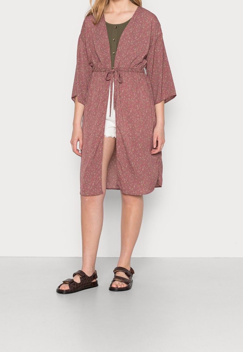Rose-colored kimono-style cover-up with floral patterns, tied at the waist. Worn over a green top and white shorts. Brown sandals complete the outfit.