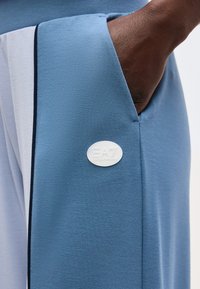 Χέρι στην τσέπη μπλε και λευκού αθλητικού παντελονιού με σήμα λογοτύπου EA7 Emporio Armani στη μπλε πλευρά.