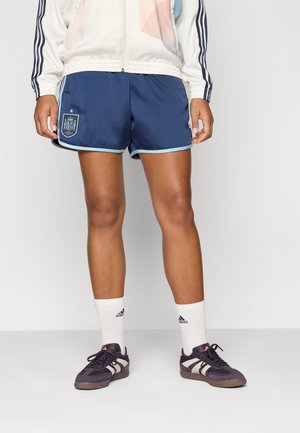 Shorts de sport navy avec des finitions bleu clair et le blason de l'Espagne, associés à des chaussettes blanches et des chaussures foncées avec des rayures et des lacets blancs.