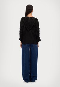 Top nero con dettaglio arricciato sul retro, maniche lunghe e chiusura a bottoni. Abbinato a jeans blu a vita alta e dallo stile morbido.