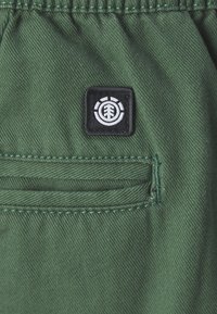 Groene stoffen shorts met een horizontale zak en een zwart vierkant label met een wit grafisch logo. De stof heeft een denim-achtige textuur.