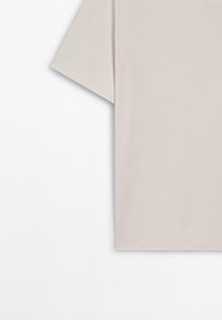 Beżowy T-shirt z krótkim rękawem pokazany od połowy klatki piersiowej do dołu, wiszący na białym tle.