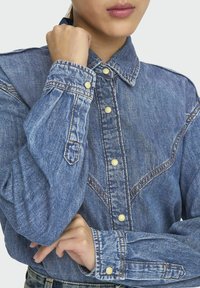 Camicia di jeans azzurra con colletto strutturato, bottoni a pressione e dettagli di cucitura evidenti. Maniche lunghe con dettagli ai polsini.