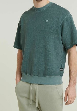 Homme portant un sweat-shirt à manches courtes bleu sarcelle délavé et un pantalon de survêtement beige clair, les mains dans les poches devant un fond uni.