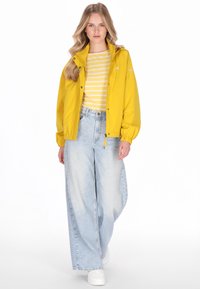 Żółta wodoodporna kurtka z suwakiem i elastycznymi mankietami, nałożona na pasiastą żółto-białą koszulę, zestawiona z jasnoniebieskimi szerokimi jeansami.