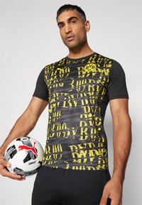 Camiseta deportiva de manga corta en negro y amarillo con estampado gráfico texturizado y escote redondo, acompañada de un balón de fútbol en blanco y negro.