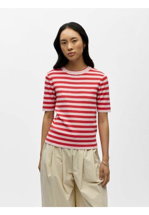 Femme aux longs cheveux foncés portant un haut à manches courtes rayé rouge et rose et un pantalon ample beige, debout devant un fond blanc uni.