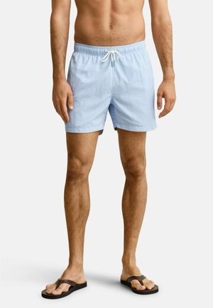 Man die lichtblauwe zwembroek met witte trekkoorden en bruine slippers draagt, staand tegen een effen achtergrond.