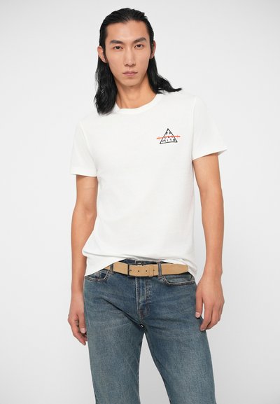 Valkoinen puuvillainen t-paita lyhyillä hihoilla ja pyöreällä kaula-aukolla, jossa on musta ja oranssi vuoristologo vasemmassa rinnassa. Yhdistetty sinisiin farkkuihin.