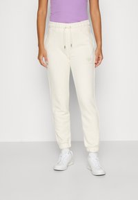 Pantaloni della tuta crema con vita elastica e polsini alla caviglia, dotati di un discreto dettaglio del logo e cordino regolabile. Abbinati a sneakers bianche.