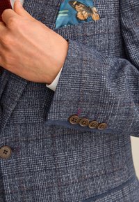 Blauer karierten Blazer aus strukturiertem Stoff, mit braunen Knöpfen und einem farbenfrohen Einstecktuch mit Schmetterlingsmotiv.