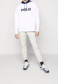 Sudadera blanca con el logotipo "POLO" en negro, capucha forrada en azul marino, combinada con pantalones cremosos ajustados y zapatillas en azul, negro y blanco.