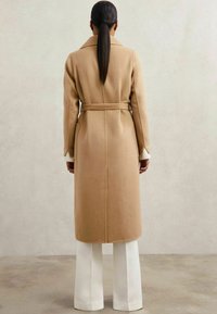 Abrigo largo color camel con cinturón en la cintura, gran collar y mangas ligeramente acampanadas, combinado con pantalones anchos blancos.