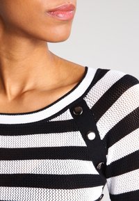 Top de punto a rayas blanco y negro con un escote amplio, detalles de botones en el hombro y una tela transpirable con textura.