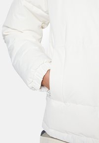 Doudoune blanche avec un design matelassé, des poignets élastiques et une poche latérale. Texture lisse avec une finition brillante.