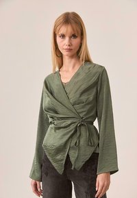 Top envolvente de color verde oliva con mangas largas, tejido texturizado y cintura anudada. Escote en V profundo y silueta relajada destacados.
