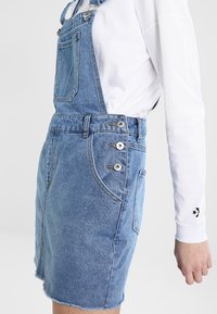 Denimöverdel i ljusblått, med justerbara remmar, metallknappar och en fransad nederkant. Sidficka synlig.