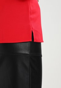 Top in tessuto rosso con fessure laterali, abbinato a una gonna in pelle nera con dettagli di cucitura visibili e una texture liscia.