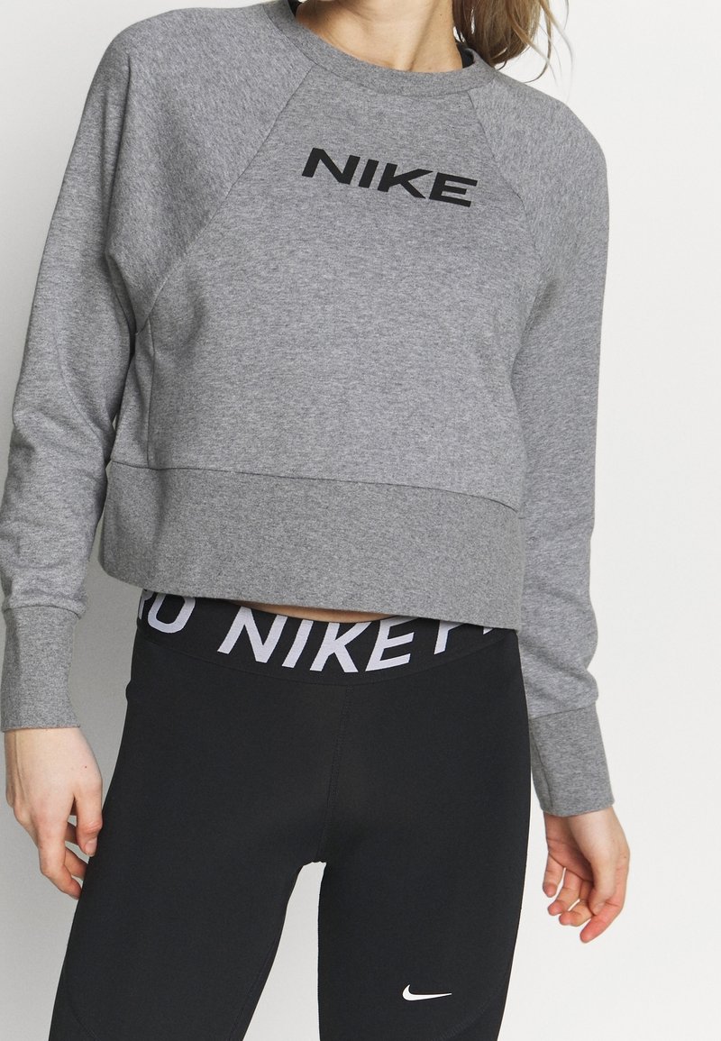 Femme portant un sweat-shirt gris Nike et des leggings noirs Nike avec un logo visible à la taille et sur la jambe, debout sur fond blanc.