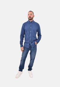 Camicia blu con bottoni realizzata in un tessuto liscio, abbinata a jeans in denim e sneakers bianche. Design semplice con colletto e logo sul petto.
