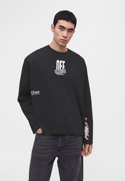 Млад мъж, носещ черна блуза с дълги ръкави Off-White и тъмносиви дънки, стои пред обикновен бял фон.