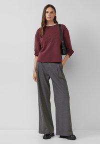 Bordeaux sweatshirt met reliëfdesign, gecombineerd met grijze wijde pantalons. Model draagt een kleine zwarte handtas. Zwarte chunky schoenen.