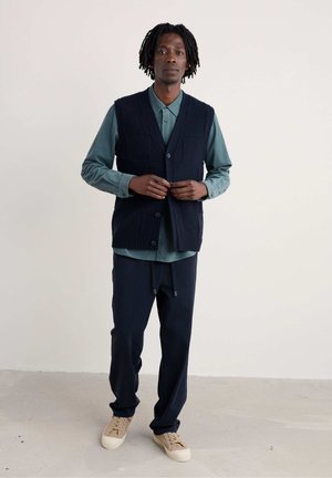 Gilet lavorato a maglia blu navy con quadrati testurizzati, indossato sopra una camicia azzurro teal; abbinato a pantaloni blu navy e scarpe beige con lacci.