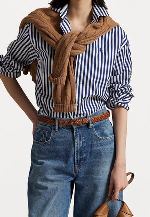 Person iført blå og hvide stribede skjorte med brun strikket sweater bundet over skuldrene, blå jeans, vævet brun bælte, holder brun håndtaske.