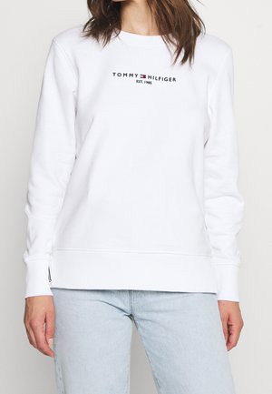 Femme portant un sweat-shirt blanc Tommy Hilfiger avec logo et un jean bleu clair, debout devant un fond uni.