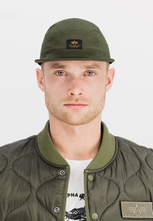 Joven con barba ligera, lleva una gorra verde y una chaqueta acolchada de color oliva sobre una camiseta blanca con diseño gráfico, mirando hacia adelante con expresión neutral.