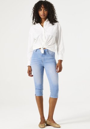 Weißes langärmeliges Hemd, das in der Taille gebunden ist, kombiniert mit hellblauen, taillenhohen Jeans mit einer engen Passform und Reißverschlussdetails am Saum.