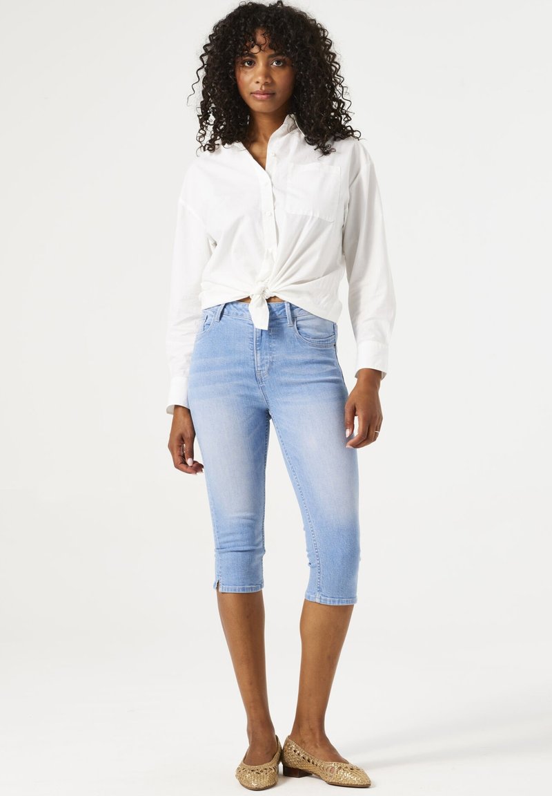 Chemise blanche à manches longues nouée à la taille, associée à un jean en denim bleu clair coupé et ajusté, avec des détails zippés à l'ourlet.
