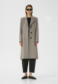 Cappotto in lana beige con colletto a dischi, tre bottoni neri e tasche laterali, indossato sopra un top e pantaloni neri, abbinato a scarpe nere.
