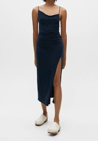 Robe midi bleu marine avec des bretelles spaghetti, tissu froncé et fente latérale, associée à des sabots beige clair avec des semelles en bois et des clous.