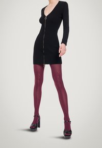 Musta ribbineuloksinen mekko, jossa on etuvetoketju ja pyöreä avainrenkaskoriste, yhdistetty viininpunaisiin sukkahousuihin, joissa on geometrinen kuvio, ja mustiin korokepohjakenkiin.