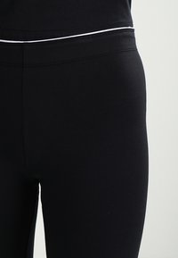 Detailaufnahme von schwarzen Leggings mit einem weißen Bund, getragen von einer Person vor einem schlichten hellen Hintergrund.