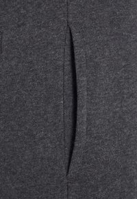 Gros plan d'une poche en tissu gris foncé avec une ouverture fendue verticale, montrant la texture et les détails de couture.