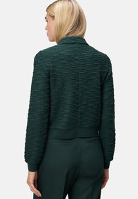Femme aux cheveux blonds portant une veste à manches longues texturée vert foncé et un pantalon assorti vert foncé, de dos face à l'appareil photo.