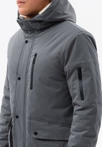 Grå vadderad jacka med hög krage, dragkedjsfickor och knäppningar. Har en texturerad utsida och kontrasterande innerfoder.