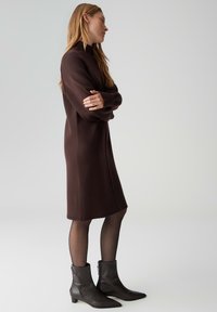 Bruine gebreide jurk met een hoge kraag, lange mouwen en een losse pasvorm. Gecombineerd met donkere enkellaarzen met een lage hak en ritsdetail.