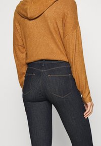 Jeans de mezclilla azul oscuro con dos bolsillos traseros y costuras naranjas, combinados con una sudadera naranja de gran tamaño que cuenta con capucha y mangas largas.